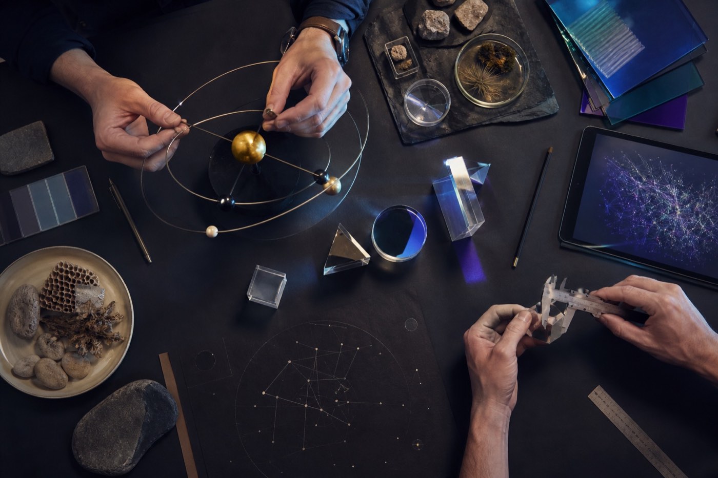 Hands use an orbital model and calipers among prisms, samples, natural materials, and a tablet on a dark worktable.